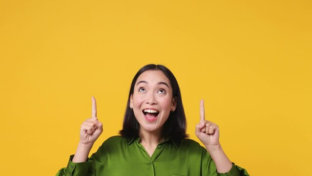 Beautiful young woman of Asian ethnicity wear green shirt pointing indicate index fingers above head on workspace area copy space mock up isolated on plain yellow color wall background studio portrait