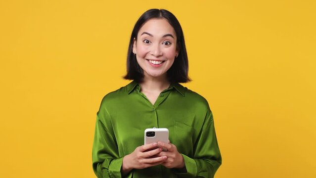 Happy smart joyful young woman of Asian ethnicity she wears green shirt hold use pointing finger on mobile cell phone just found out great big win news isolated on plain yellow color wall background
