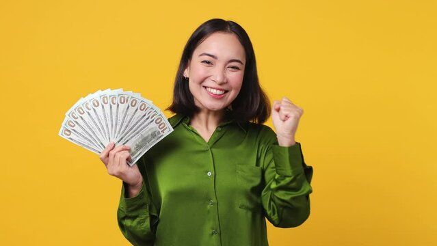 Young woman of Asian ethnicity 20s she wear green shirt hold in hand fan of cash money in dollar banknotes doing winner gesture celebrate isolated on plain yellow color wall background studio portrait