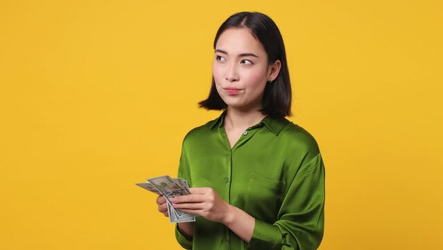 Side view young accountant woman of Asian ethnicity 20s she wear green shirt hold in hands count stack of cash money in dollar banknotes isolated on plain yellow color wall background studio portrait