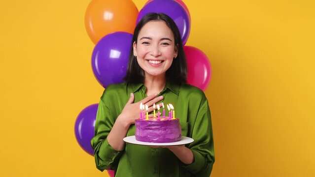 Happy young woman 20s she wear green shirt celebrate birthday holiday party with bunch of colorful air balloons hold cake make wish blow burning candles isolated on plain yellow color wall background