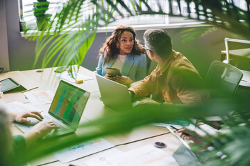 Cheerful diverse coworkers sitting at table with laptop and working on project in office