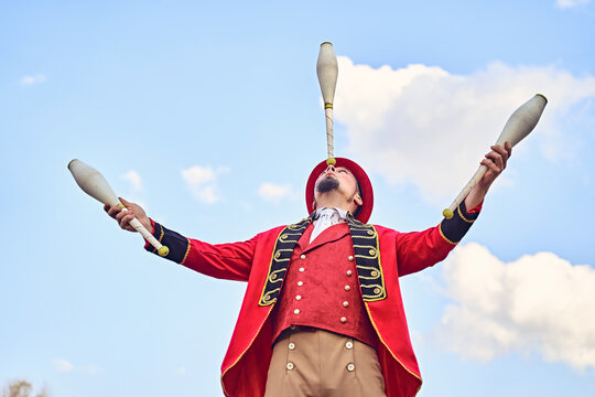 From below man in red costume balancing club on nose while juggling against cloudy blue sky during performance in park
