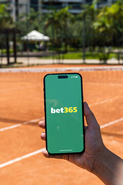 Girl holding an iPhone 14 Pro smartphone with Bet365 betting provider app on screen. Clay tennis court in the background. Rio de Janeiro, RJ, Brazil. November 2022