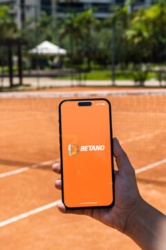 Girl holding an iPhone 14 Pro smartphone with Betano betting provider app on screen. Clay tennis court in the background. Rio de Janeiro, RJ, Brazil. November 2022