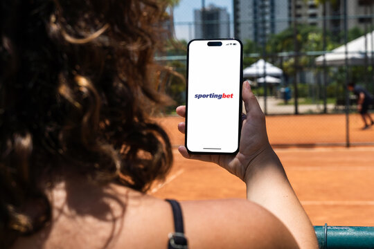 Girl holding an iPhone 14 Pro smartphone with Sportingbet betting provider app on screen. Clay tennis court in the background. Rio de Janeiro, RJ, Brazil. November 2022