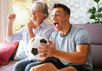 Cheerful couple of young boy and senior grandmother soccer fans watching a football game on tv...