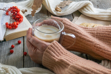 On a wooden table there is a mug of hot coffee with foam held by women's hands in a warm sweater, cookies, a book, a branch of mountain ash and a warm scarf. The concept of warmth, comfort
