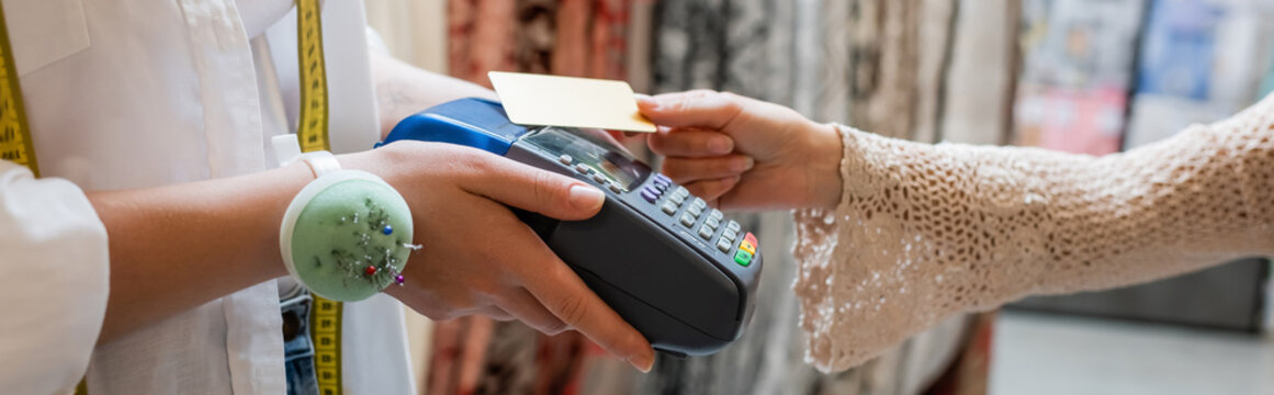cropped view of seller with needle cushion on hand holding credit card reader near customer in textile shop, banner