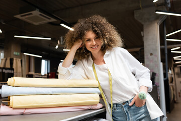 happy saleswoman with needle cushion posing with hand in pocket while leaning on desk with colorful fabric rolls