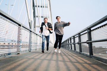 Two happy young people man and woman in casual clothes running across the footbridge in city.