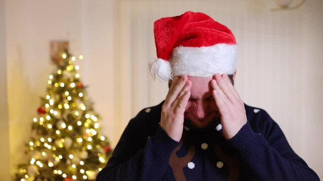 holiday anxiety depression mental health during Christmas celebration with family, close up of shocked bearded male wearing santa hat with xmas tree lighted up in background