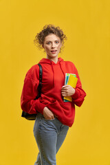 Front view of pretty girl standing, walking, looking at camera. Young student with curly hair holding books, hand in pocket, smiling with open mouth. Concept of modern culture.