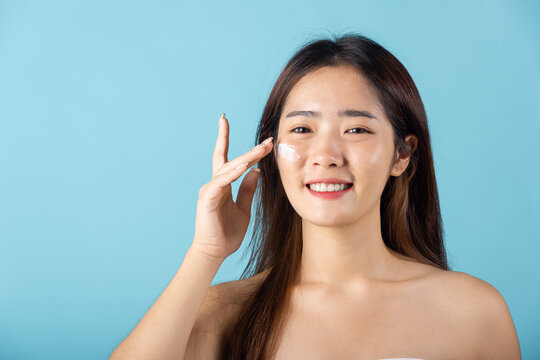 Skin care and beauty concept. young woman applying moisturizer cream on her face studio shot isolated on blue background, Happy beauty portrait female gets cream on her cheek, health clean skin