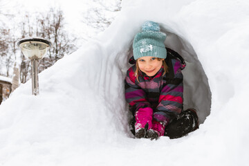 Fototapeta na wymiar Happy little girl sitting in a snow cave made in a snowdrift looking at camera smiling