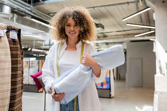 Joyful Saleswoman With Measuring Tape Carrying Blue Fabric Roll In Textile Shop