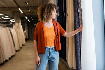 Side view of smiling curly woman looking at curtains in textile shop - obrazy, fototapety, plakaty