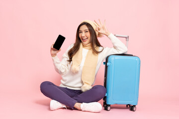 Happy Asian woman traveler sitting on floor with luggage and holding mobile phone isolated on pink...