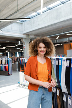 joyful woman looking at camera near variety of multicolored fabric in textile shop