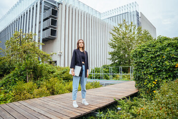 Full height portrait of female student with laptop on modern building terrace with green garden.