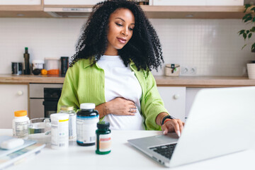 Picture of adorable african american pregnant woman ordering prenatal supplements in web store sitting at kitchen table with bottles of vitamins, scrolling on laptop. Healthy pregnancy - obrazy, fototapety, plakaty