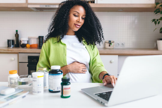Picture of adorable african american pregnant woman ordering prenatal supplements in web store sitting at kitchen table with bottles of vitamins, scrolling on laptop. Healthy pregnancy