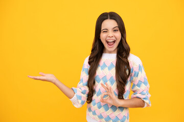Close up portrait of teenager child girl showing at copy space, pointing to ads advertising, isolated over yellow background. Mock up copy space. Excited face. Amazed expression, cheerful and glad.