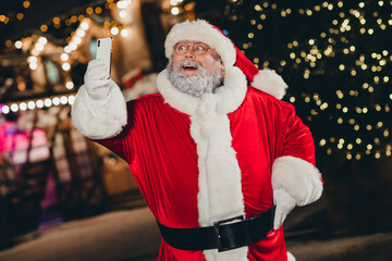 Portrait of funky granddad santa hold telephone make selfie record video newyear tree lights city center outdoors