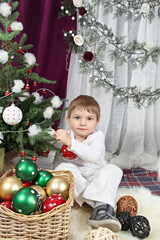 Fototapeta na wymiar Portrait of a cute smiling 3 year old boy in white clothes decorating a Christmas tree with colorful balls, New Year's interior