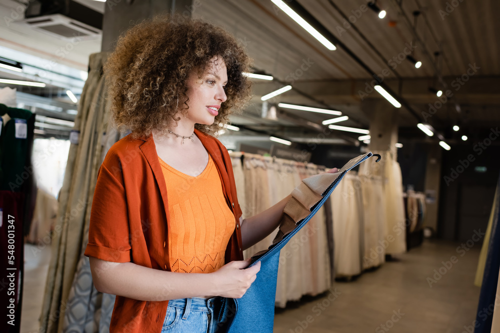 Wall mural young smiling woman holding fabric samples in blurred textile shop