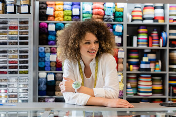 curly saleswoman with measuring tape and needle cushion looking away near counter and rack with needlework accessories