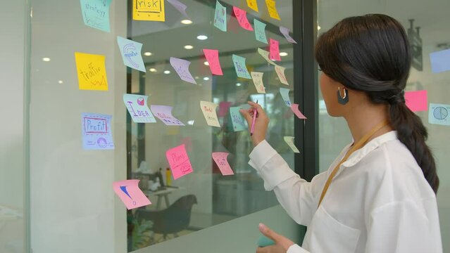 Asian businesswoman thinking and planning  at office and use post it notes to share idea. analyzing concept. Startup office. Sticky note on glass wall.