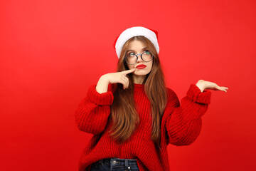 A young woman in a santa hat on a red background makes a difficult choice, an empty space for an...