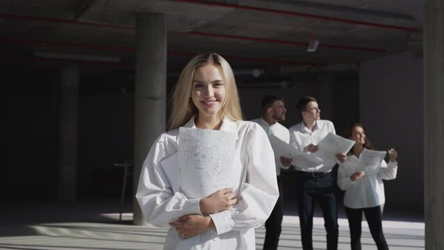 Positive blonde woman looks in camera smiling at building construction site against colleagues discussing project plan on background. Portrait of happy young female investor holding paper drawing
