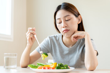 Obraz na płótnie Canvas Diet in bored face, unhappy beautiful asian young woman, girl on dieting, holding fork in salad plate, dislike or tired with eat fresh vegetables. Nutrition of clean, healthy food good taste.