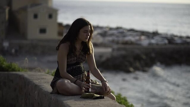 Young Spanish woman drinks a beer at sunset sitting on the edge of the seacoast.