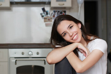 Fototapeta na wymiar A beautiful young brunette girl looks directly into the camera and smiles with a wide snow-white smile. The kitchen is visible in the background