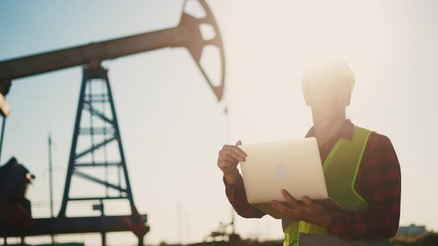 oil industry. engineer next to an oil rig. oil business production finance concept. gas production. a worker with a laptop monitors oil production next to an station. sun a worker in hard hat works