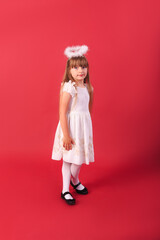 A little blue-eyed girl in a white angel dress against a red background. Studio shot of a baby girl in an angel dress.