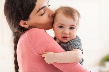 Caring young mother holding and kissing sweet little baby girl, mom and adorable kid bonding at home, copy space