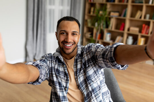 Excited arab guy taking selfie and smiling at mobile phone camera, sitting in armchair at home, free space
