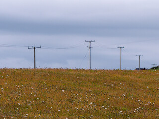 A pole of power lines in a field under the sky. Minimalistic landscape. Black electric post on grass field under white cloudy sky