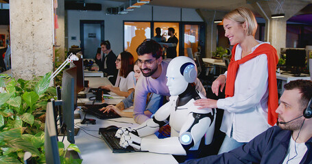 Happy multietnic people standing and looking at humanoid working in office. Robot typing on...