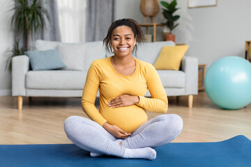 Cheerful pretty young black pregnant lady with big belly practicing yoga on mat in modern living room interior