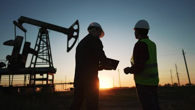 oil production. two workers a work next to an oil pump at sunset silhouette. industry business oil production lifestyle concept. engineers in hardhats studying oil production figures