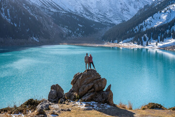 Romantic couple standing on a huge boulder on a vantage point over stunning turquoise lake surrounded by snowy mountains on a sunny day. Pressing to each other.