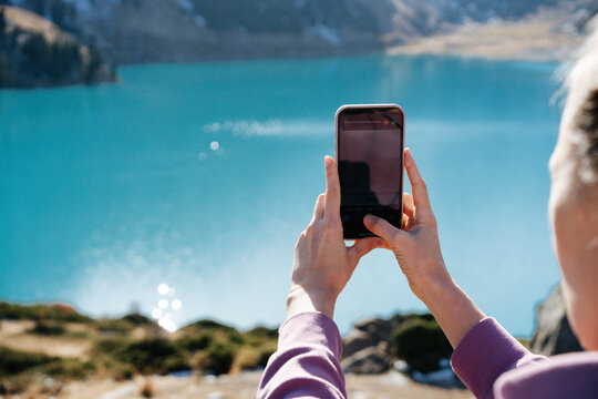 Over the shoulder image of a woman taking a picture with her phone through polaroid sunglasses of the stunningly beautiful lake surrounded by snowy mountains.