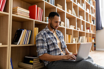 Young arab guy studying online from home, using laptop pc, sitting on floor near bookshelves, copy...