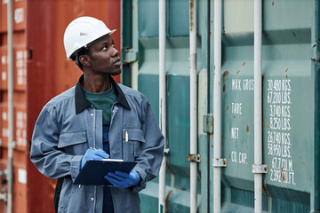 Waist up portrait of young black man wearing hardhat while working at shipping dock and holding...