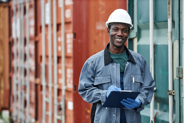 Waist up portrait of smiling young worker wearing hardhat while posing in container shipping docks, copy space